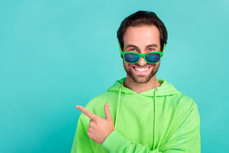 Photo of positive smart guy indicate forefinger empty space wear sunglass green hoodie isolated teal...