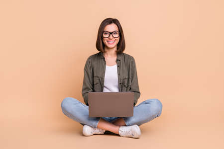 Photo of nice confident smart lady sit floor hold laptop wear glasses khaki shirt jeans isolated beige color backgroundの写真素材