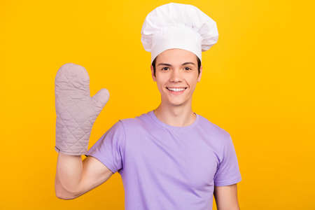 Portrait of attractive cheerful guy wearing chef headwear gloves waving hi isolated over bright yellow color backgroundの写真素材