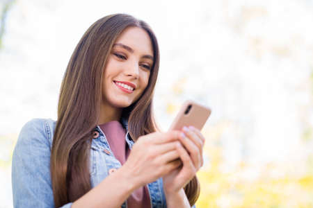 Photo of sweet shiny woman wear denim jacket smiling walking typing modern device outdoors urban city parkの写真素材