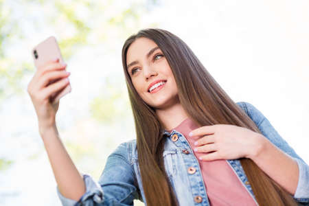 Low angle view portrait of attractive cheerful girl using device taking making selfie blogging outdoor strolling fresh open airの写真素材