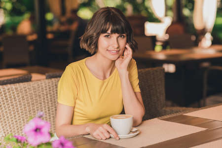 Photo of dreamy charming young lady dressed yellow t-shirt sitting cafeteria drinking beverage smiling outdoors urban parkの写真素材