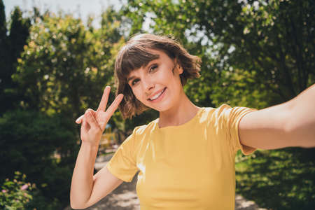 Photo of pretty adorable young woman wear yellow t-shirt smiling recording video showing v-sign outside city streetの写真素材