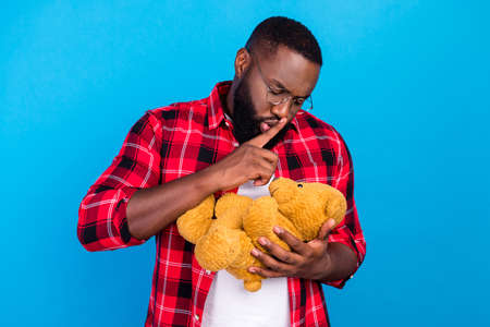 Photo of serious young afro american man hold hands teddy bear silent mute finger lips isolated on blue color backgroundの写真素材