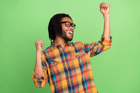 Photo of dreamy lucky dark skin guy dressed checkered shirt glasses dancing rising fists smiling isolated green color backgroundの写真素材
