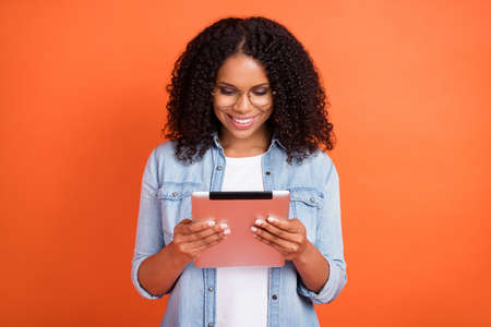Photo of funny young brunette lady look tablet wear jeans shirt eyewear isolated on orange color...