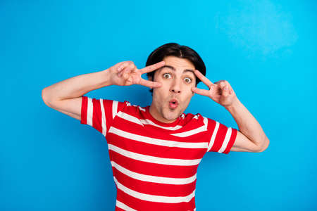 Photo of cute shocked young man wear striped t-shirt showing v-signs cover eyes isolated blue color backgroundの写真素材