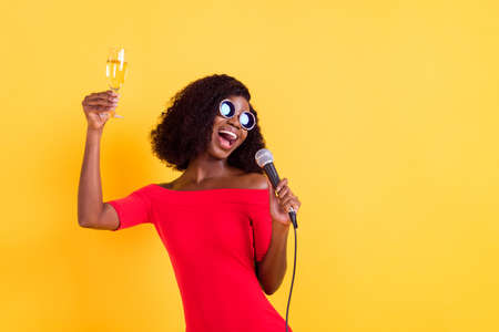 Photo of young happy crazy happy afro girl singing karaoke hold champagne in glass isolated on yellow color backgroundの写真素材