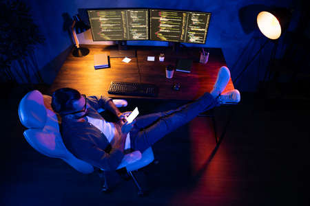 Full body above high view of young guy sit put legs on desktop texting communicate lamp light late...