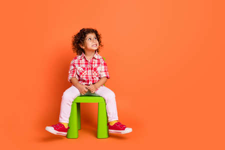 Photo of dreamy sweet schoolgirl wear plaid outfit smiling looking empty space sitting green chair isolated orange color backgroundの写真素材