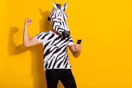 Photo of excited freak guy blogger in zebra mask use smart phone fist up win followers post isolated over yellow color backgroundの写真素材