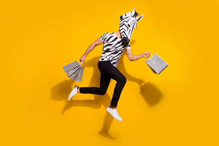 Photo of cheerful hurrying man wear zebra print t-shirt mask holding shoppers running fast jumping high isolated yellow color backgroundの写真素材