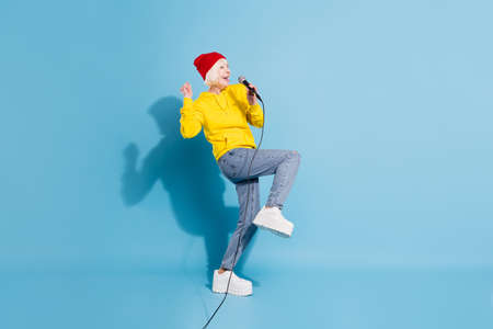 Photo of adorable lucky age woman dressed yellow sweatshirt red headwear rising fist singing karaoke isolated blue color backgroundの写真素材