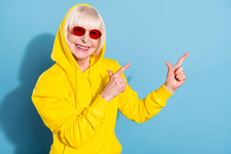 Photo of sweet nice pretty old woman point fingers empty space advert wear sunglass hoodie isolated on blue color backgroundの写真素材
