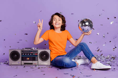 Portrait of handsome trendy cheerful boy holding disco ball having fun showing v-sign isolated over...