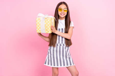 Photo portrait of small schoolgirl in overall glasses smiling keeping present box isolated pastel...