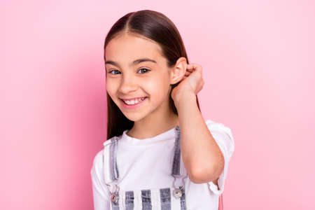 Photo portrait of small schoolgirl smiling touching hair in overall isolated on pastel pink color...