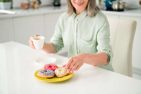 Cropped view portrait of attractive elderly retired pensioner cheerful grey-haired woman drinking coffee eating bakery at home light white indoorsの写真素材