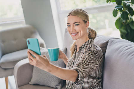 Photo of positive domestic mature lady sit divan hold mug latte make selfie smart phone in living roomの写真素材