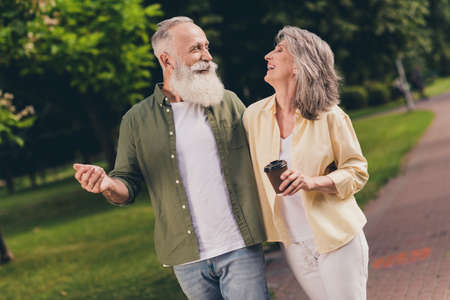 Photo of charming positive pensioner friends dressed casual shirts smiling drinking tea embracing walking outdoors urban city parkの写真素材