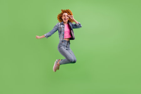 Full length photo of charming funny young lady dressed denim clothes glasses smiling jumping high isolated green color backgroundの写真素材