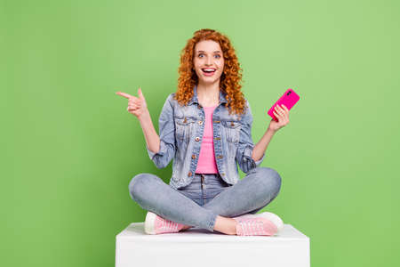 Full size photo of young girl happy smile use cellphone point finger empty space ad promo suggest isolated over green color backgroundの写真素材