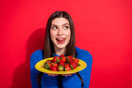 Photo of dreamy young woman look empty space hold hands plate berry isolated on red color backgroundの写真素材