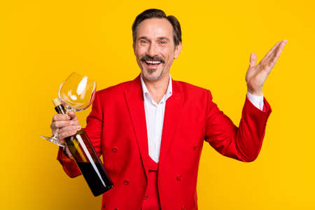 Photo of smiling man in red color costume hold wine bottle celebrate party anniversary isolated on yellow color backgroundの写真素材