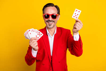 Portrait of cheerful good mood croupier hold poker card choose pick wear red suit isolated on yellow color backgroundの写真素材