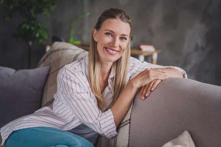 Photo of cute young blond lady sit on sofa wear white shirt in free-time apartment room aloneの写真素材