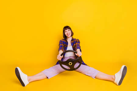 Portrait of attractive cheerful girl sitting holding in hands steering wheel having fun isolated over bright yellow color backgroundの写真素材