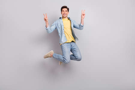 Photo of pretty cheerful guy dressed denim shirt showing two v-signs jumping high isolated grey...