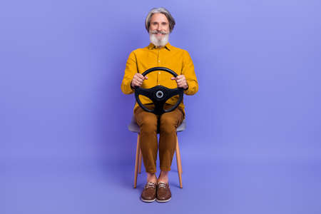 Portrait of attractive cheerful gray-haired man sitting riding steering wheel isolated over bright violet purple color backgroundの写真素材