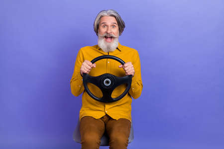 Portrait of nice amazed cheerful gray-haired man riding steering wheel having fun isolated over bright violet purple color backgroundの写真素材