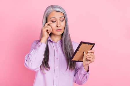 Portrait of attractive sad sullen gray-haired woman looking at photo crying isolated over pink pastel color backgroundの写真素材
