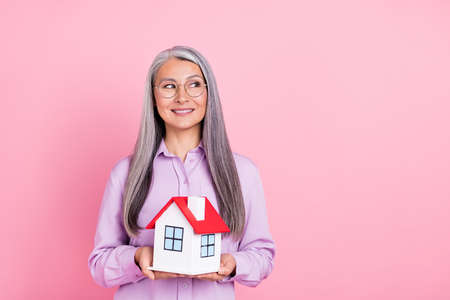 Portrait of attractive gray-haired cheery minded woman holding house thinking copy space isolated over pink pastel color backgroundの写真素材