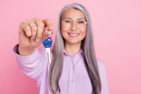 Portrait of attractive cheerful gray-haired woman giving room hotel house key isolated over pink pastel color backgroundの写真素材