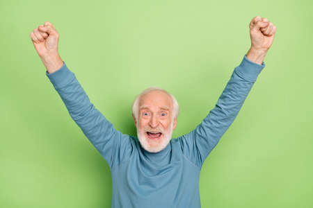 Photo of delighted astonished man raise fists celebrate goal accomplishment isolated on green color backgroundの写真素材