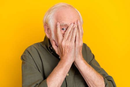 Portrait of scared aged person arms palms covering face one eye peek look camera isolated on yellow color backgroundの写真素材