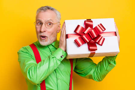 Photo of aged man happy positive smile curious shake gift box birthday anniversary isolated over yellow color backgroundの写真素材