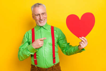 Photo of aged man happy positive smile point finger paper heart advert suggest promo isolated over yellow color backgroundの写真素材