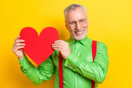 Photo portrait old man smiling in spectacles showing red heart shaped postcard on date isolated vivid yellow color backgroundの写真素材
