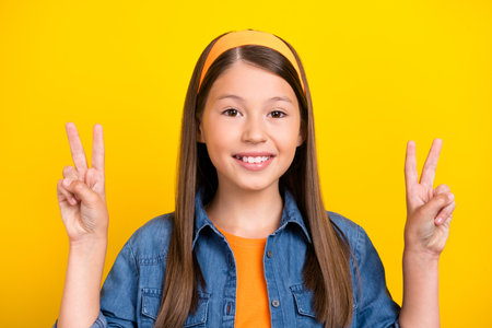 Photo of funny charming preteen girl dressed jeans shirt showing two v-signs smiling isolated yellow...