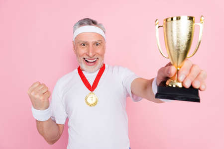 Photo portrait of senior man in headband smiling showing prize gesturing like winner champion isolated pastel pink color backgroundの写真素材