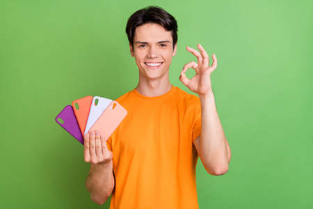 Photo of young happy charming man make okay sign hold phone case isolated on green color backgroundの写真素材