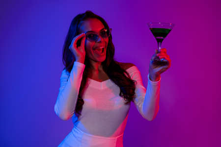 Photo of hooray young brunette lady drink alcohol wearing white dress spectacles isolated over neon colorful backgroundの写真素材