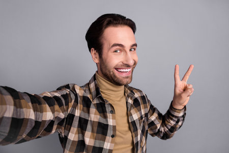 Photo of excited funny guy dressed checkered shirt tacking selfie showing v-sign isolated grey color...