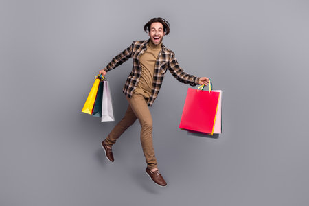 Photo of pretty impressed man wear plaid shirt jumping running holding bargains isolated grey color...