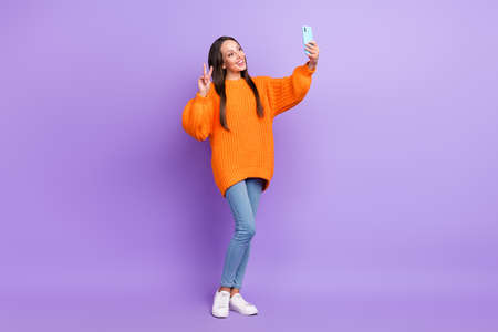 Photo of friendly cute lady hold phone make selfie show v-sign wear orange sweater isolated purple...