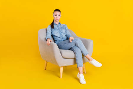 Photo of serious confident young woman wear denim shirt glasses sitting chair isolated yellow color backgroundの写真素材